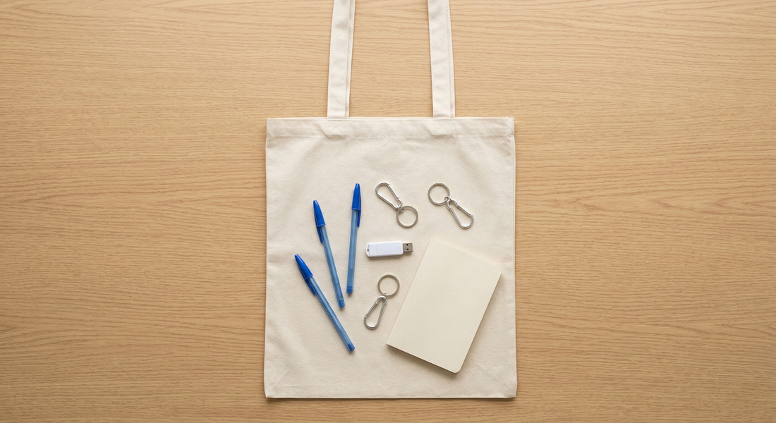 Comparativa de merchandising: Tote Bag de 180g cruda con bolígrafos y llaveros sobre mesa de madera.